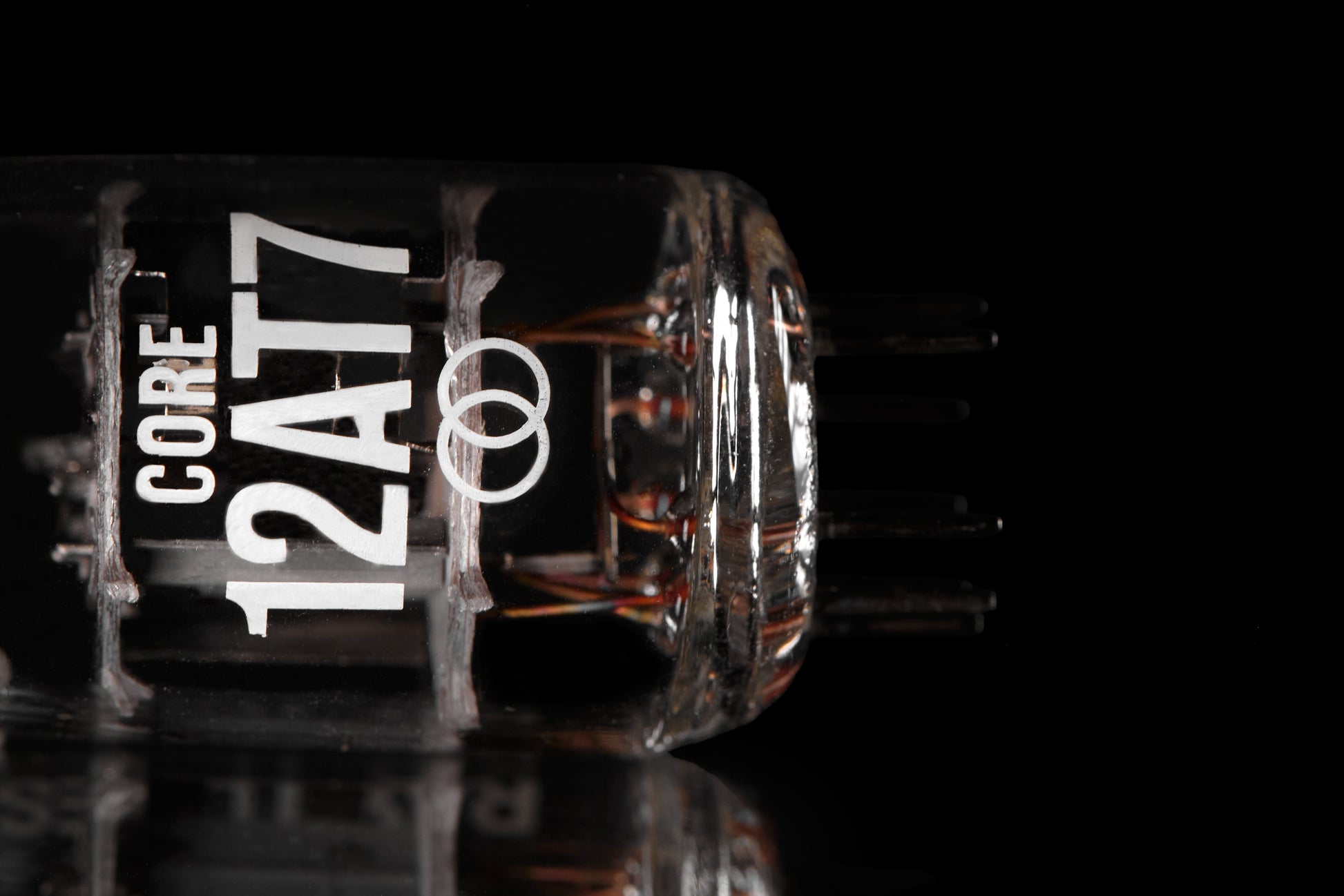 A close-up studio shot of a clear glass vacuum tube labeled 'CORE 12AT7' in white text, with internal copper filaments visible, set against a black background with a reflective surface below.