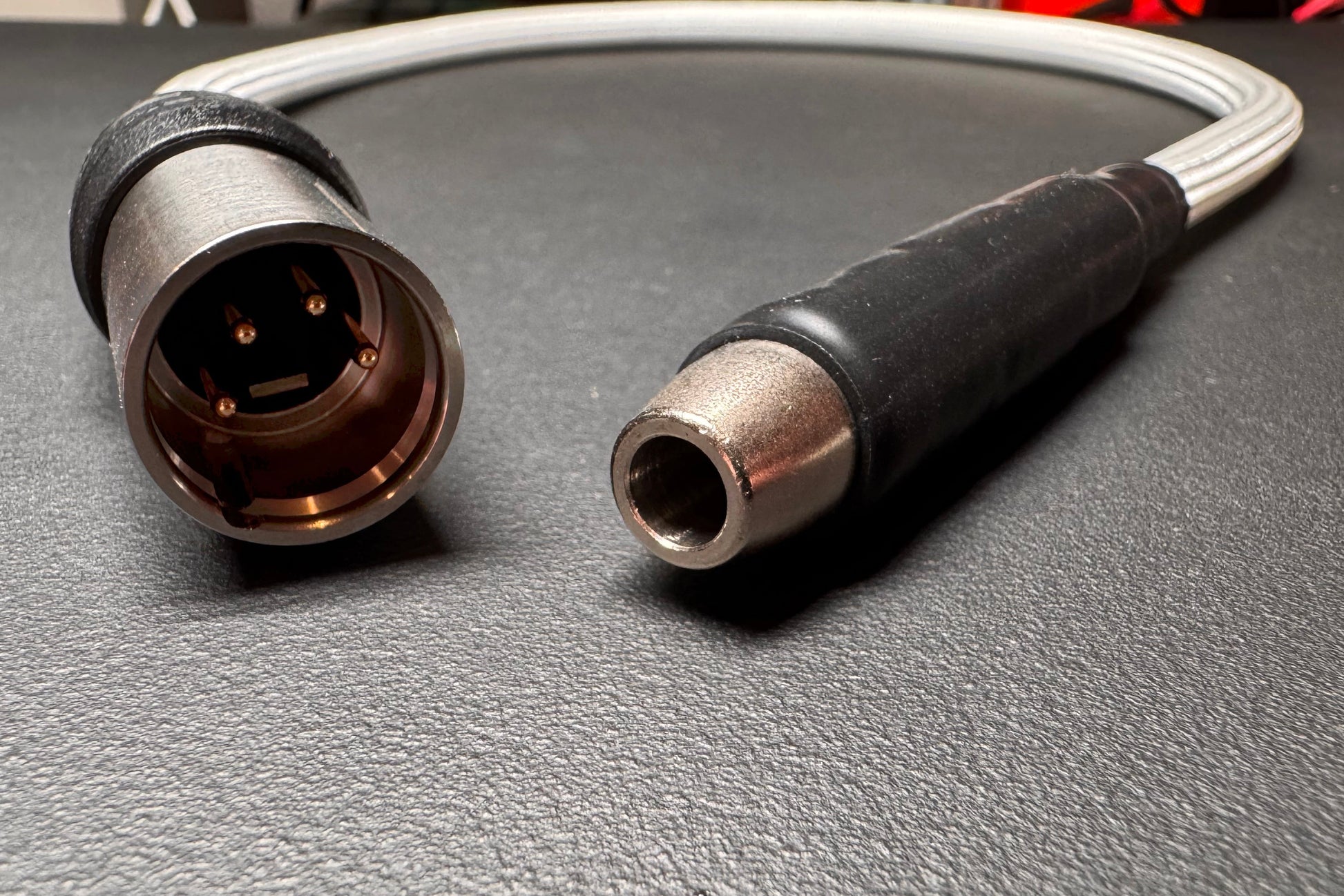 A close-up of a cable connector showing both ends on a dark textured surface. One end has a metallic multi-pin socket with a black protective ring, and the other has a metal plug with a black grip sleeve.