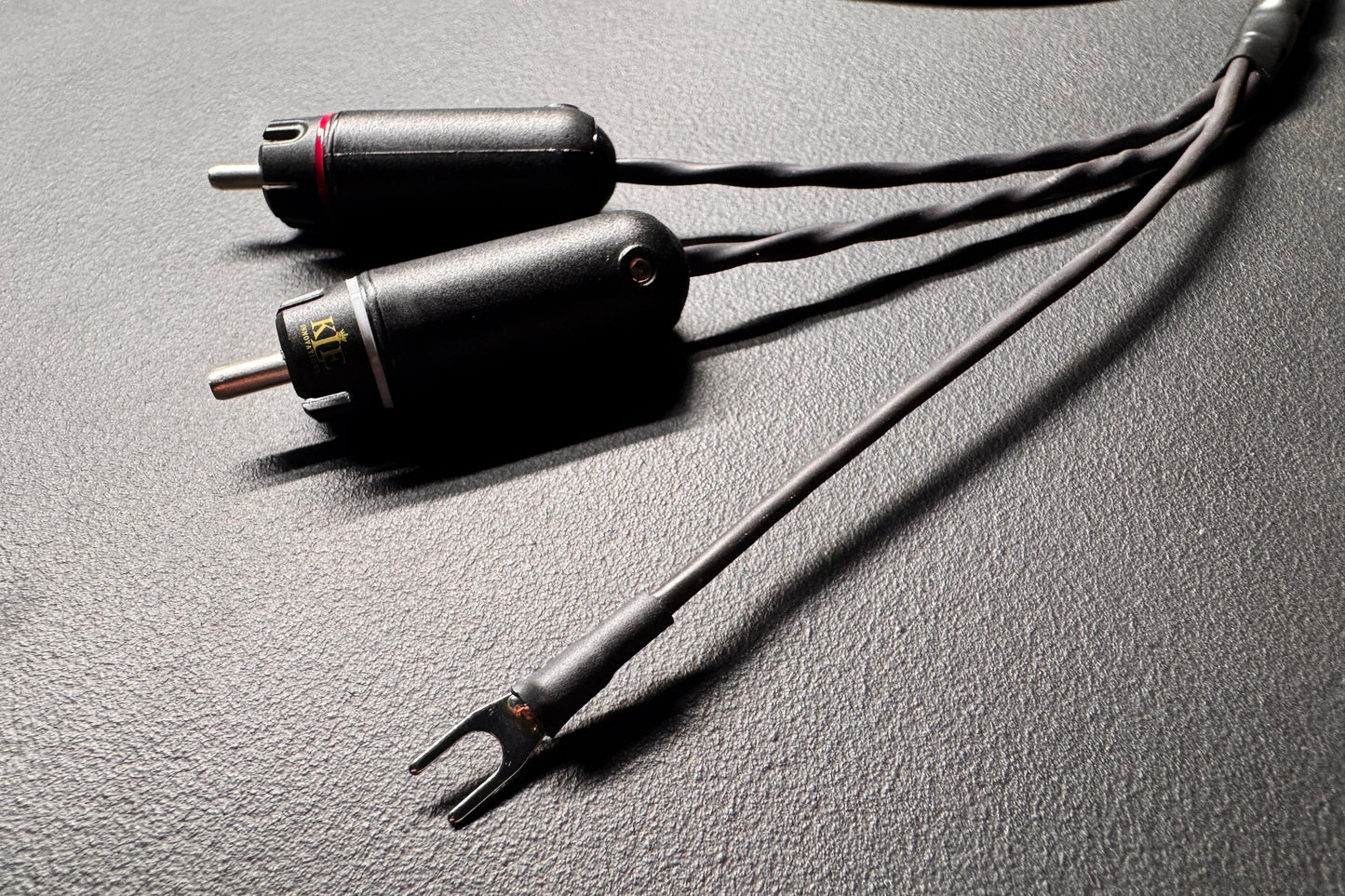 A close-up of a black audio adapter cable with two cylindrical connectors and a Y-shaped split plug, resting on a textured dark gray surface.