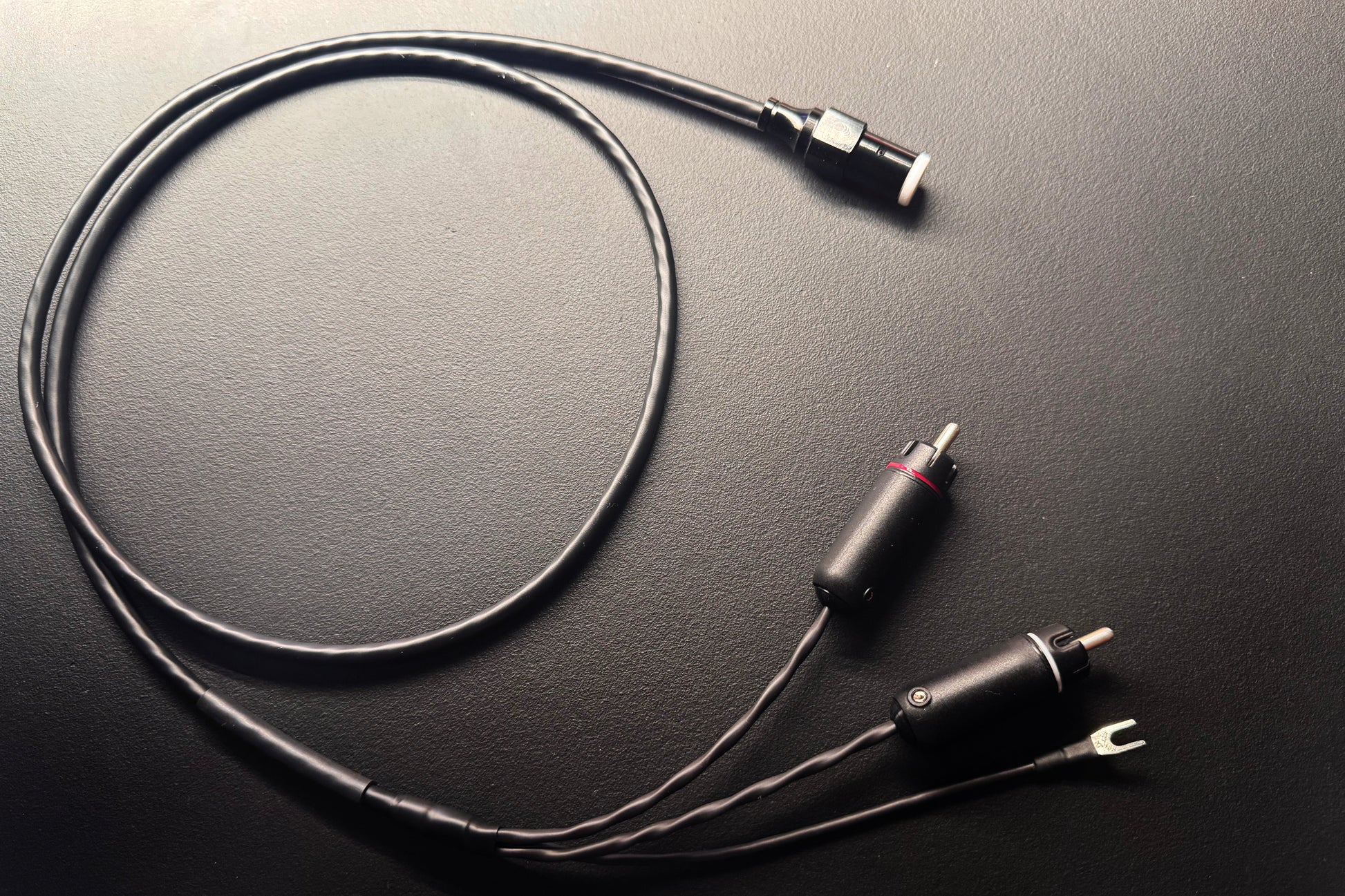 A black audio adapter cable with a 3.5mm jack, a 2-pin connector with a red ring, and a 2-pin terminal clip, displayed on a dark textured surface.