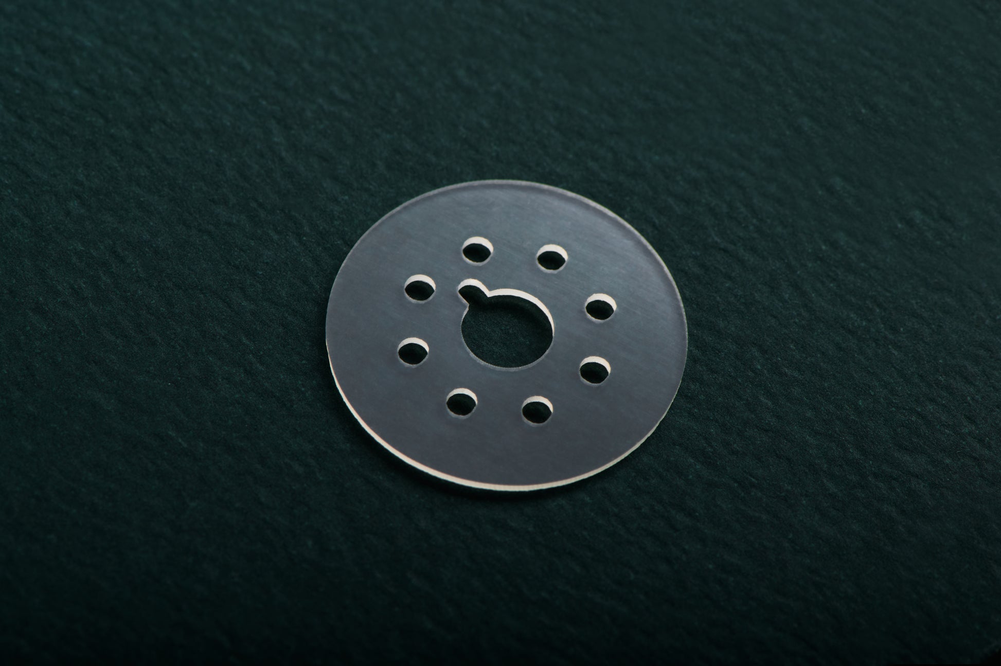 A small round metallic disc with multiple circular cutouts, photographed on a dark textured background.