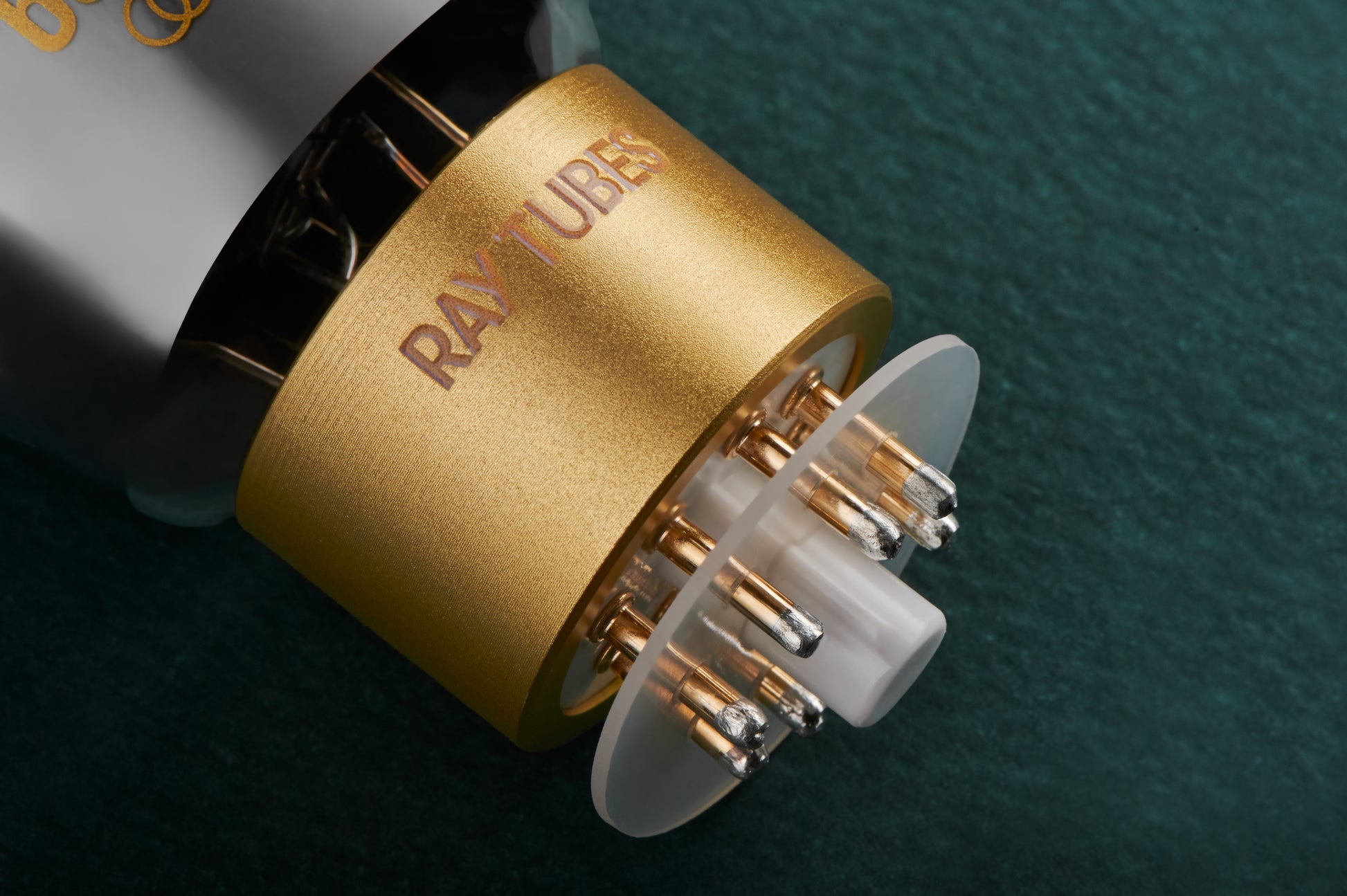 Close-up of a Ray-Tubes electronic component with a brushed gold cylindrical body, multiple metal pins, and a translucent white circular element, set against a dark green textured background.