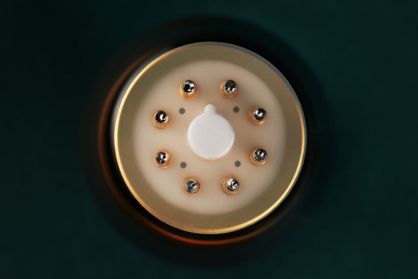 A close-up top-down view of a circular beige and gold device with a central white button and small metallic details, set against a dark teal background.