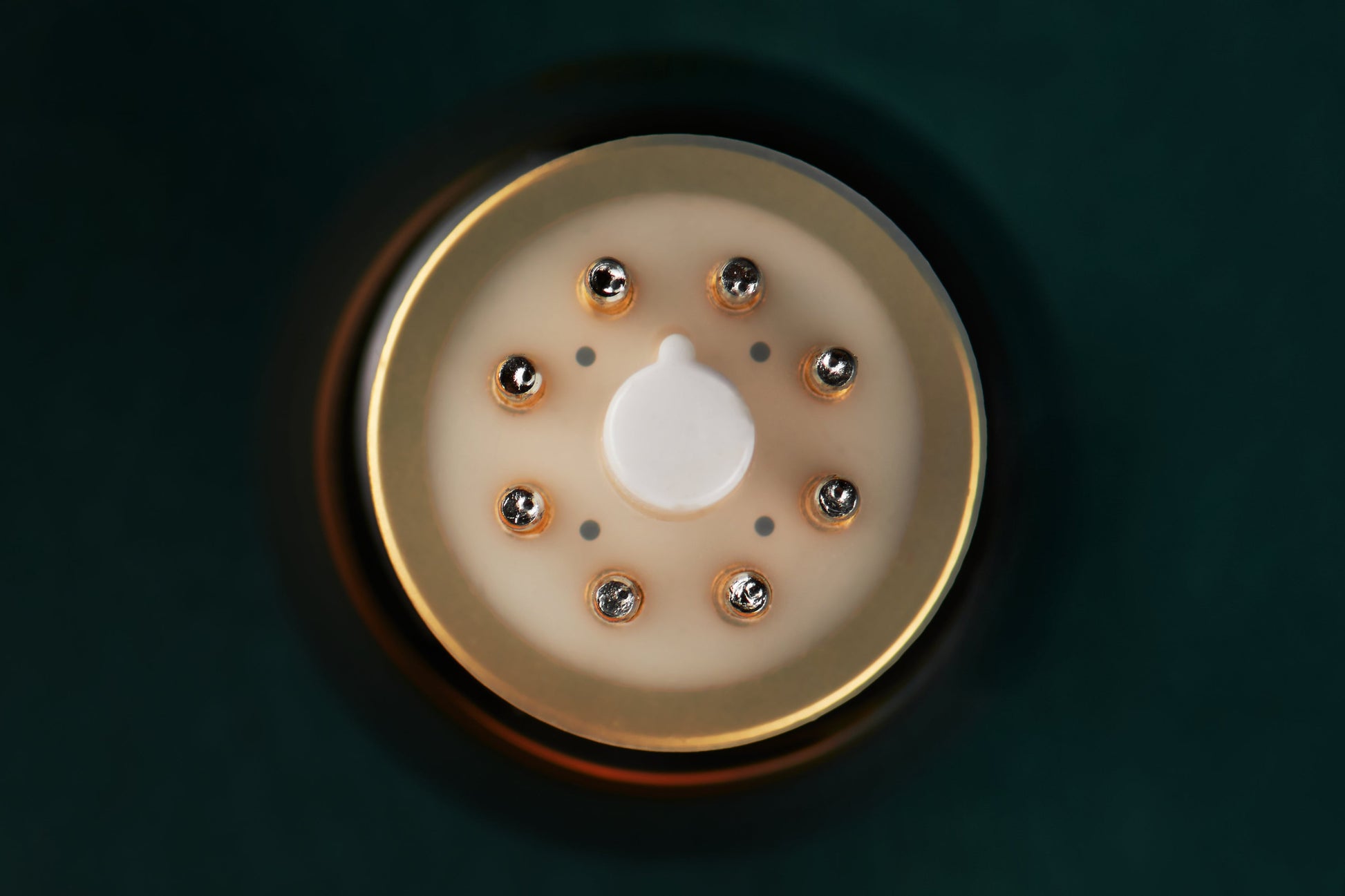 A close-up top-down view of a circular beige and gold device with a central white button and small metallic details, set against a dark teal background.