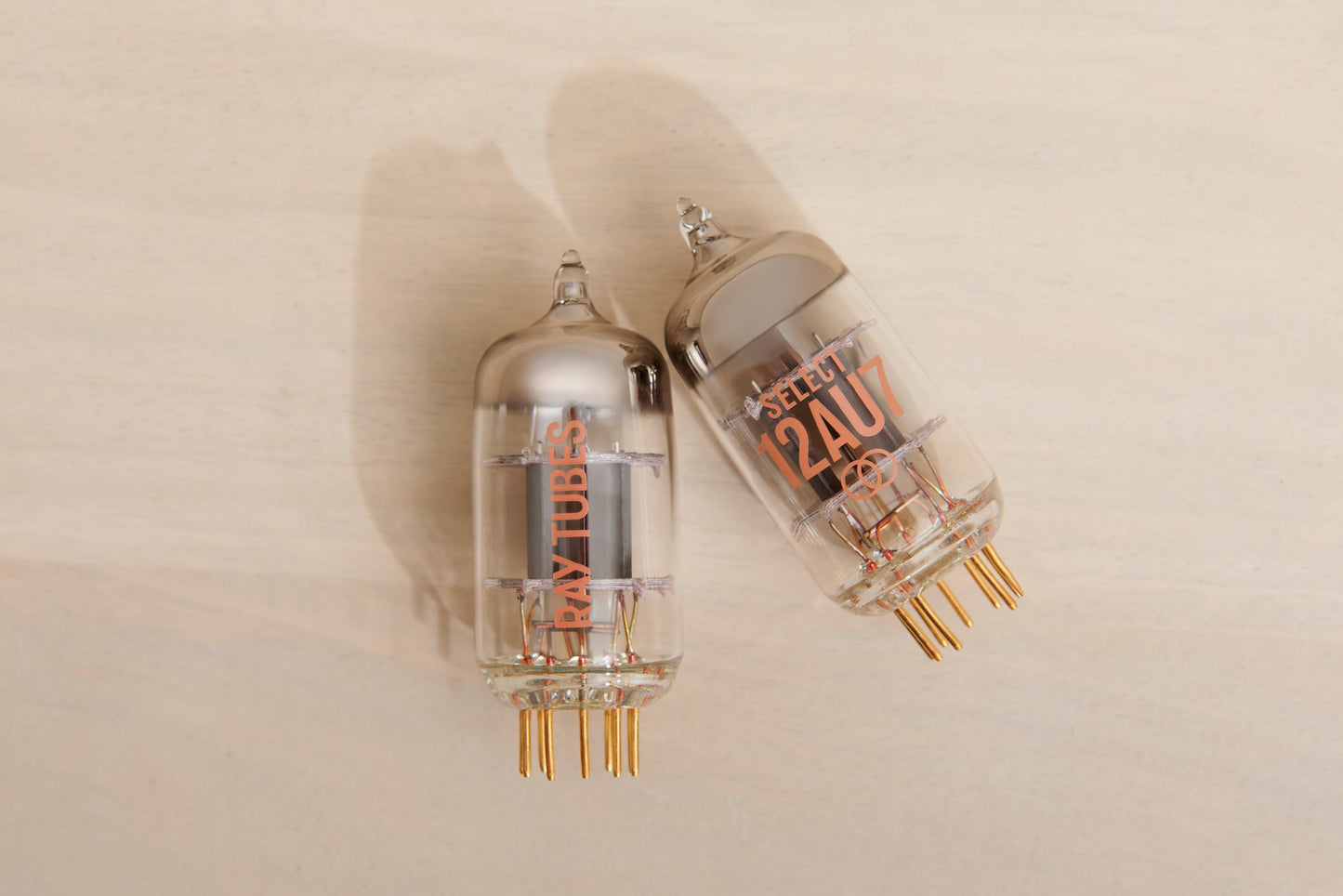 Two glass vacuum tubes, one labeled 'Ray Tubes and the other 'SELECT 12AU7', resting on a light beige wooden surface.