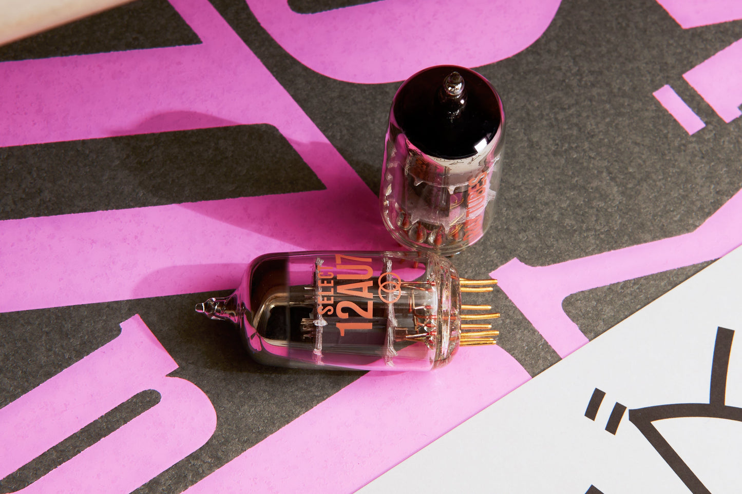 Two glass vacuum tubes, one labeled 'SELECT 12AU7' with gold pins, resting on a pink and black graphic background.