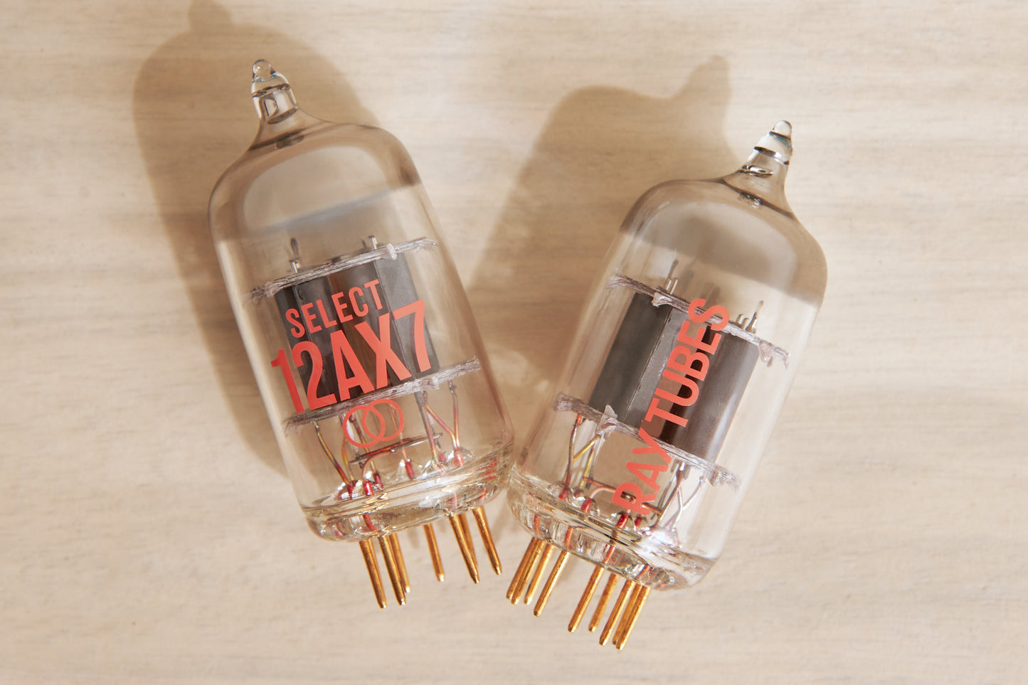 Two glass vacuum tubes with copper pins, featuring red printed text 'SELECT 12AX7' and 'RAY TUBES', displayed on a light wood background.