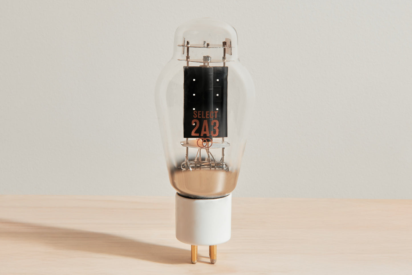 A vintage-style vacuum tube lamp with a clear glass body, internal black and copper components, and a white base, standing on a light wood surface against a neutral beige background.