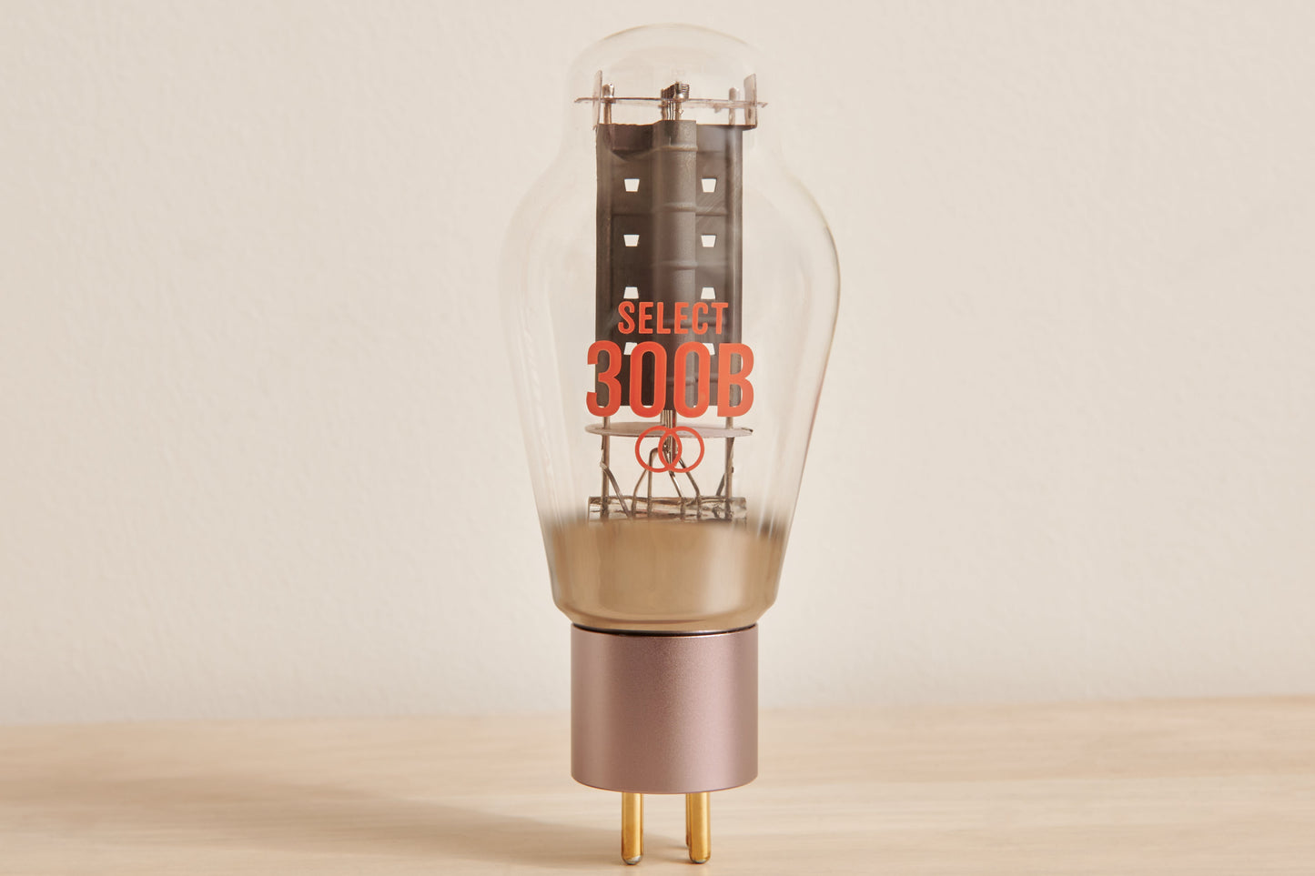 A Select 300B glass vacuum tube lamp with a clear bulb, orange printed branding, and a metallic rose gold base on a light wood surface against a neutral beige background.