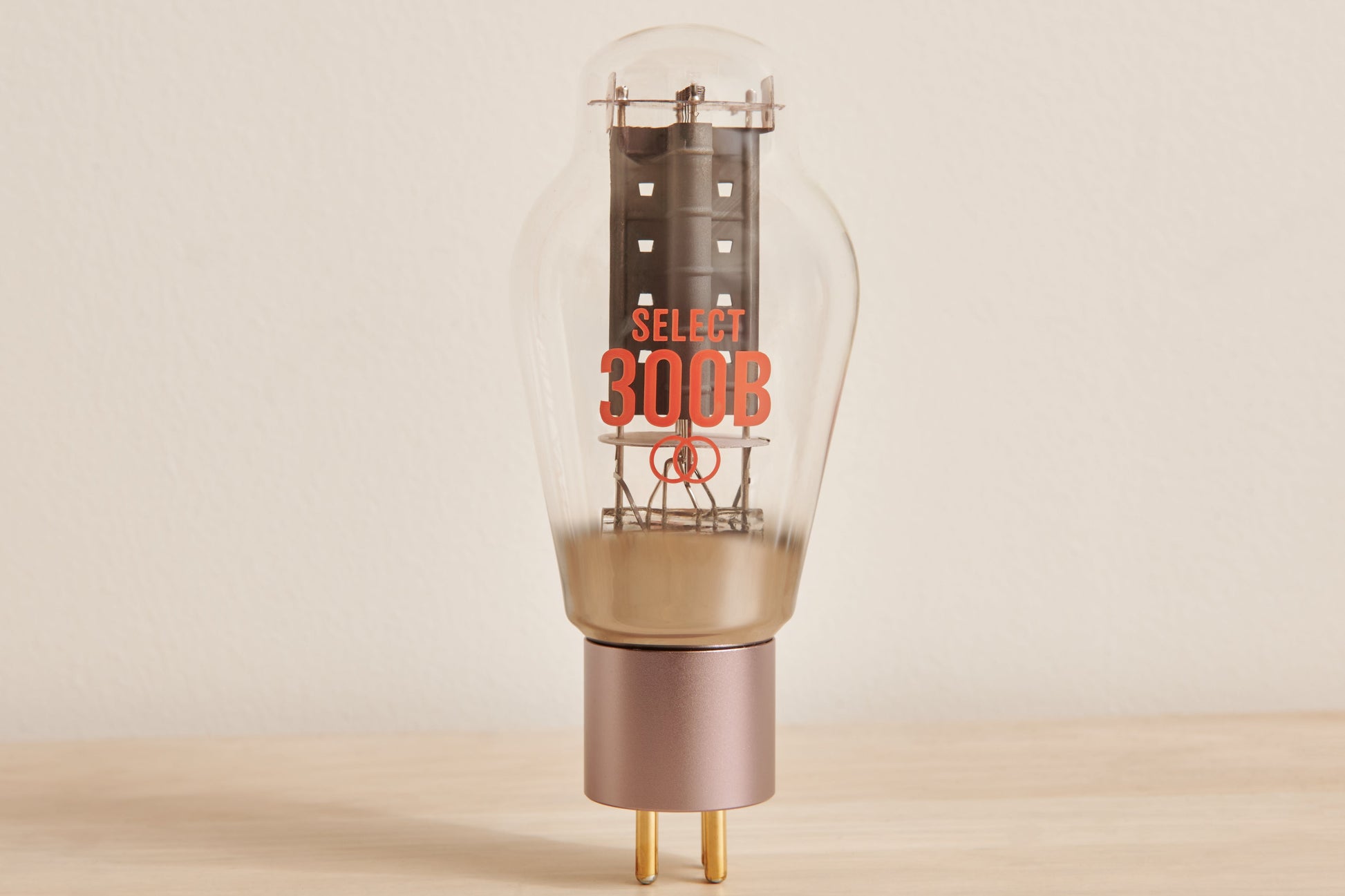 A Select 300B glass vacuum tube lamp with a clear bulb, orange printed branding, and a metallic rose gold base on a light wood surface against a neutral beige background.
