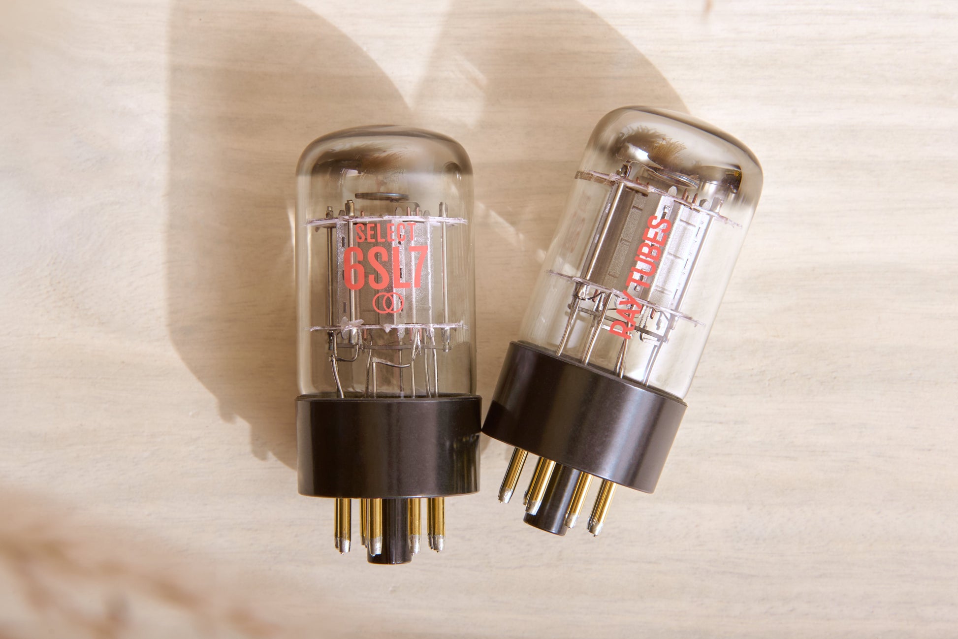 Two glass vacuum tubes with black bases and gold pins, standing upright on a light wood surface. One tube has red printed text reading 'SELECT 6SL7', and the other shows internal red elements.