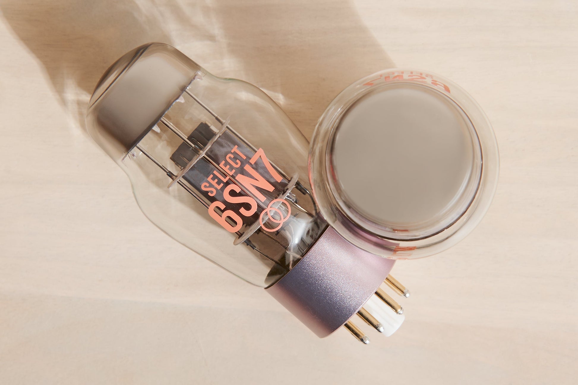 A glass vacuum tube component with metallic internal elements and a lavender-purple base, shown alongside a circular clear cap or cover on a light wood surface. The tube has visible text reading 'SELECT 6SN7'.