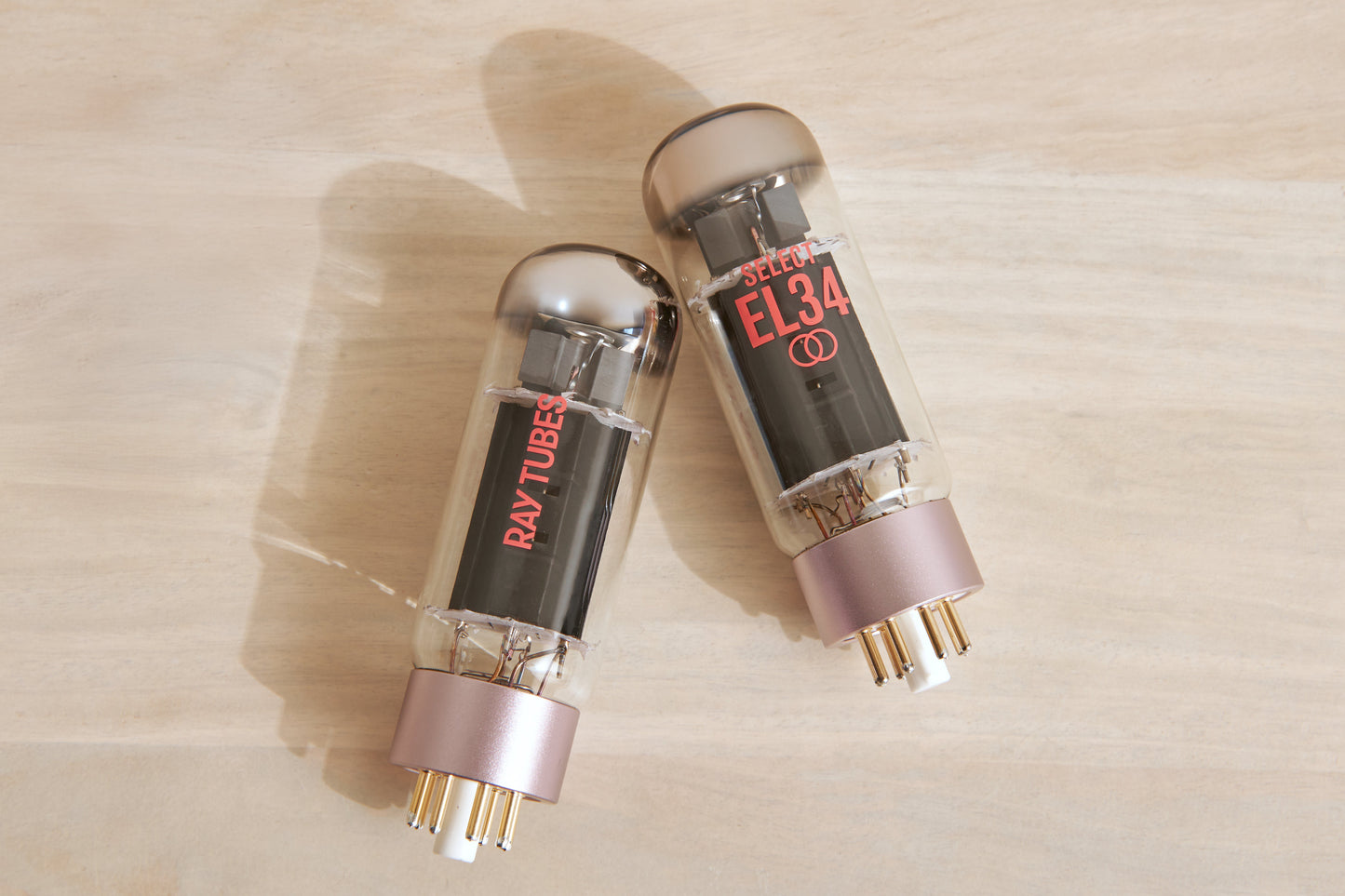 Two Ray-Tubes EL34 audio power tubes with rose gold bases and gold pins, resting on a light wood surface.