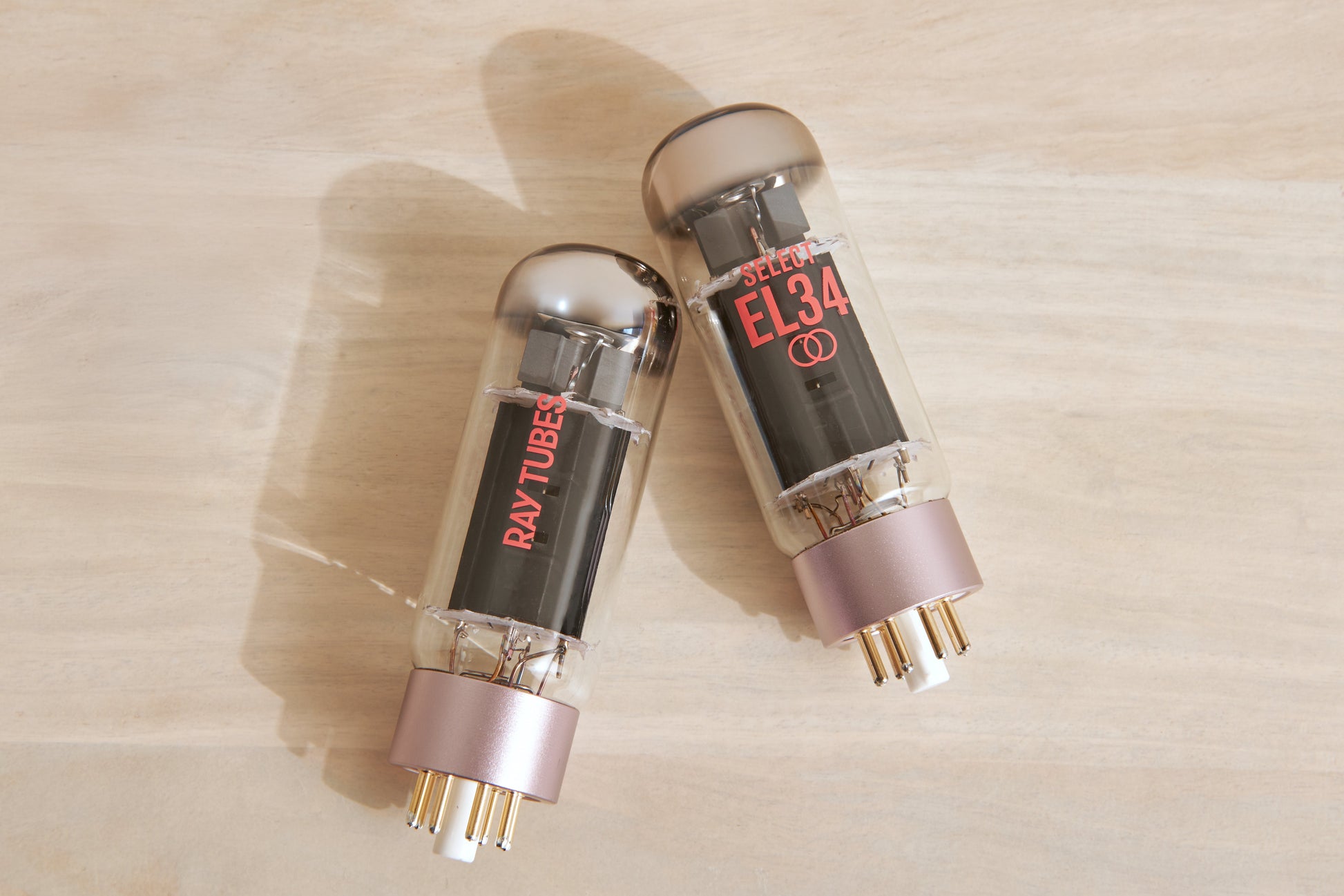 Two Ray-Tubes EL34 audio power tubes with rose gold bases and gold pins, resting on a light wood surface.