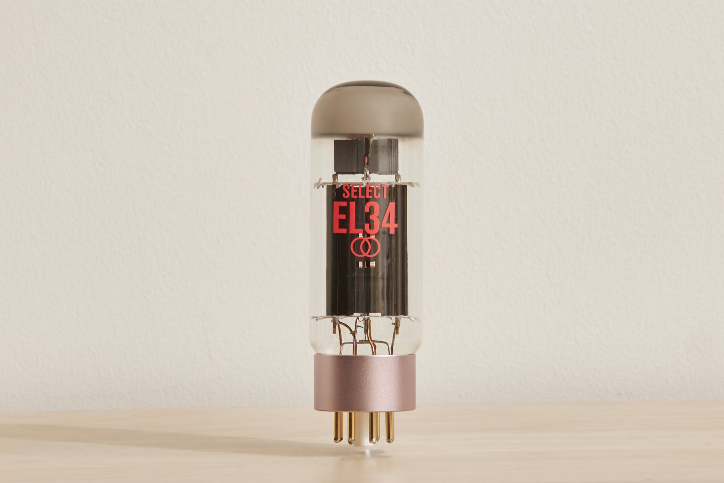 A single EL34 vacuum tube standing upright on a light wood surface against a plain off-white wall. The tube has a clear glass body, a dark gray top cap, a pink metallic base, and red printed EL34 text on the front.