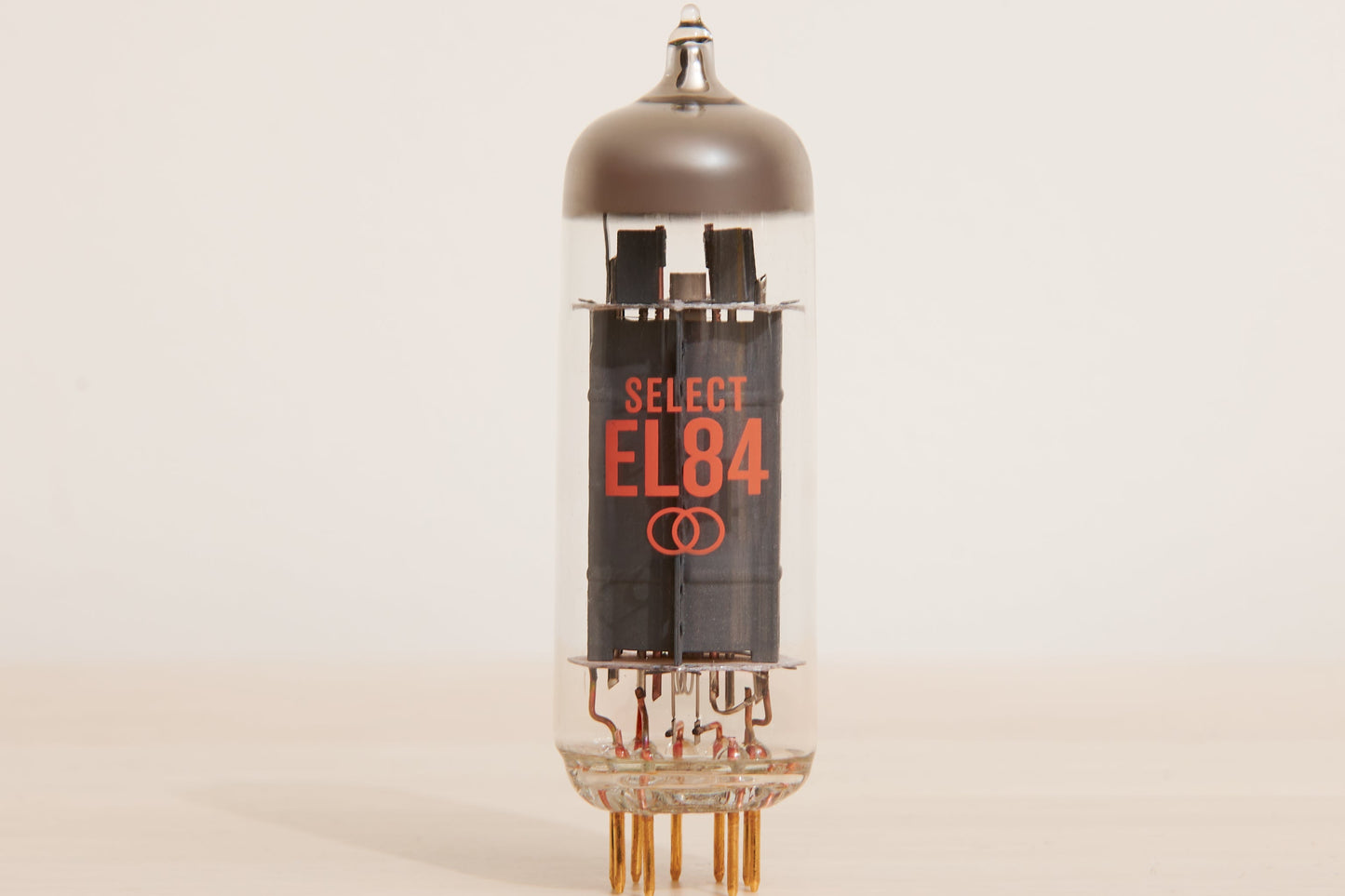 A vintage Select EL84 vacuum tube with a clear glass body, black internal components, and orange printed text, standing on a light beige surface against a neutral off-white background.