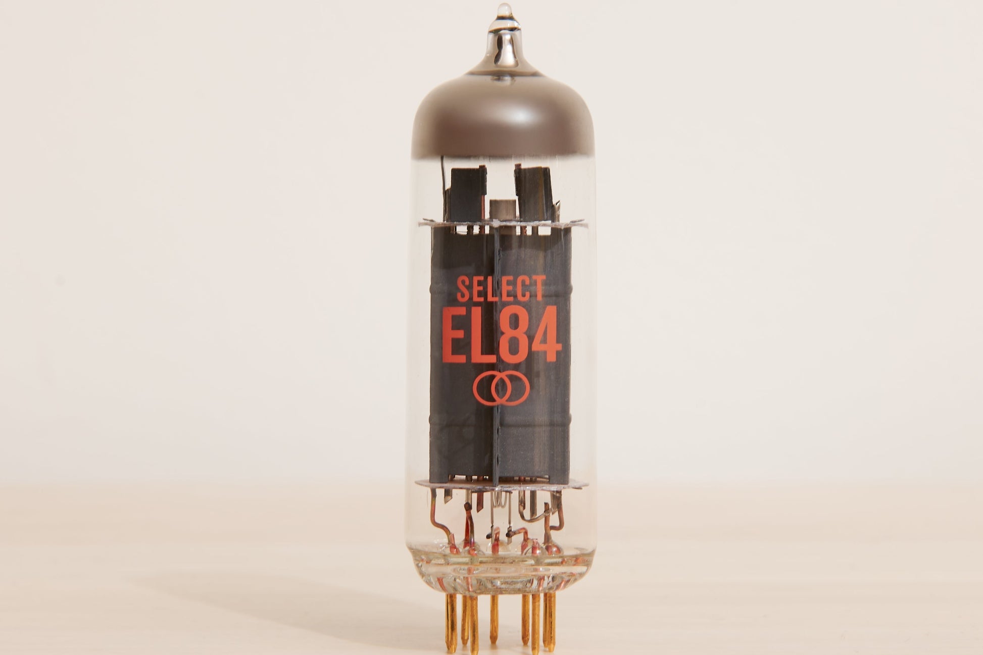 A vintage Select EL84 vacuum tube with a clear glass body, black internal components, and orange printed text, standing on a light beige surface against a neutral off-white background.