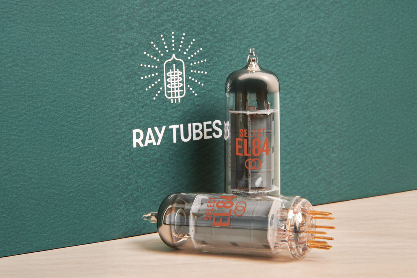 Two glass ray tube electronic vacuum tubes displayed on a light wood surface against a dark teal textured background. One tube stands upright with orange printed text reading 'SELECT EL84', and the other lies on its side showing multiple metal pins.