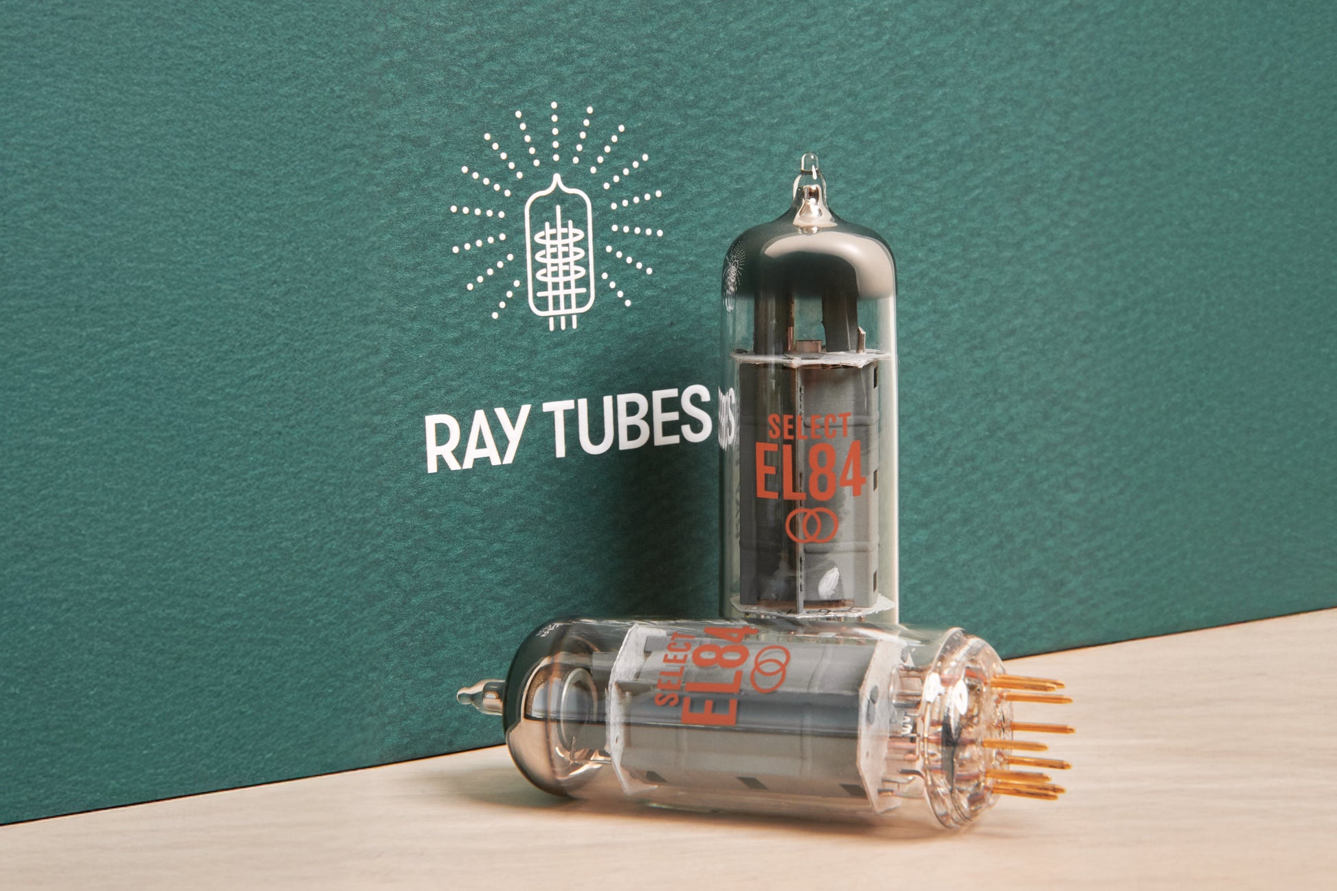 Two glass ray tube electronic vacuum tubes displayed on a light wood surface against a dark teal textured background. One tube stands upright with orange printed text reading 'SELECT EL84', and the other lies on its side showing multiple metal pins.